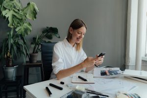 A female professional checks her smartphone while working at a desk with architectural plans.