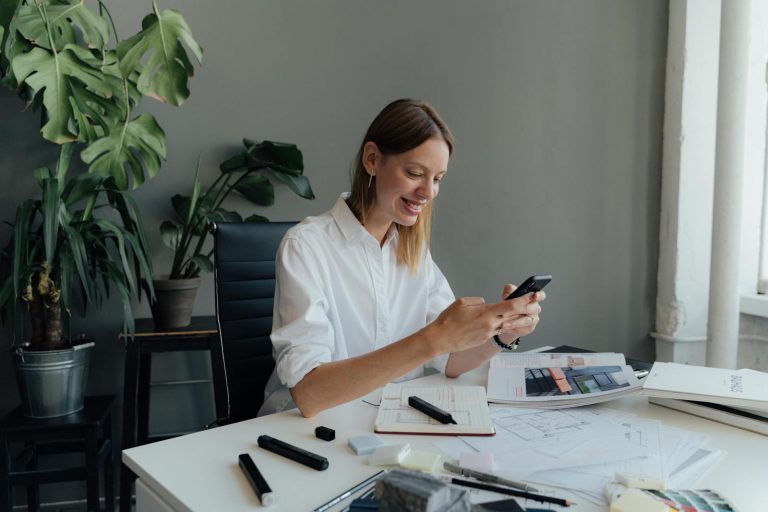 A female professional checks her smartphone while working at a desk with architectural plans.